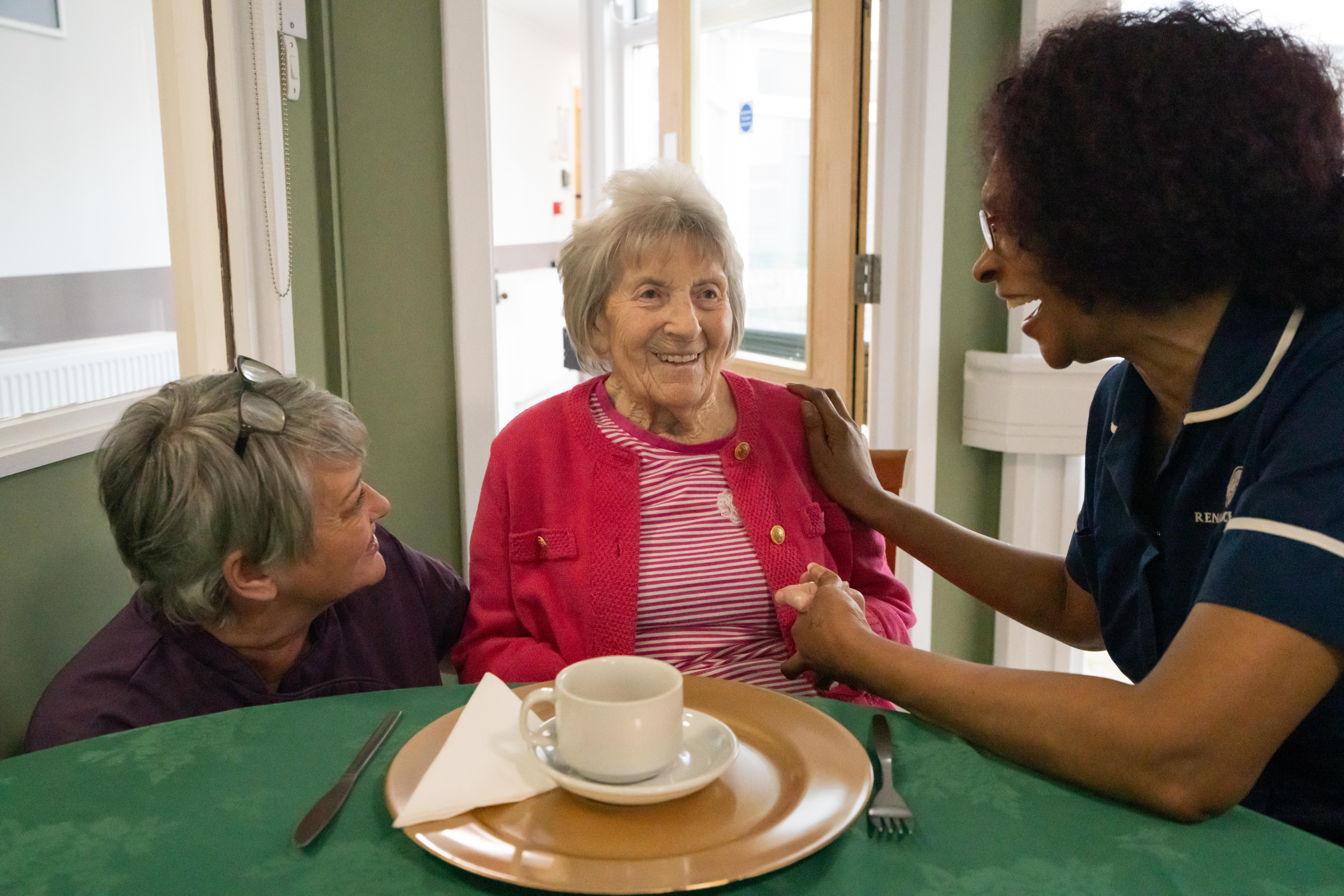 A woman in a pink cardigan sits at a table with a green tablecloth, enjoying a cup of tea. She is sat by two caregivers who are laughing and talking with her. One caregiver, in a navy blue uniform, gently holds the resident’s hand, creating a warm and social atmosphere.