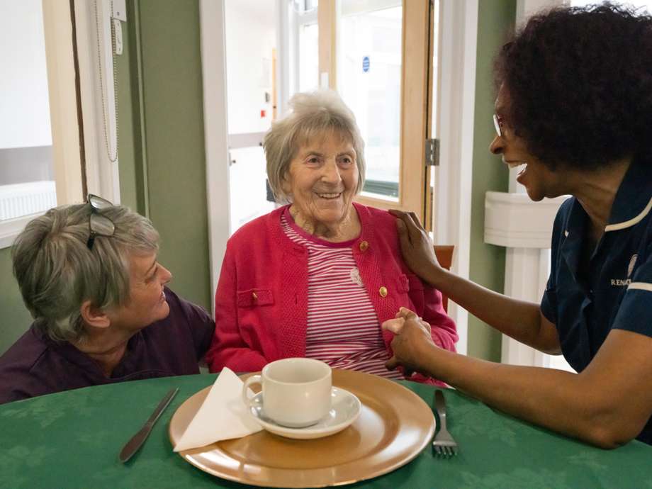 A woman in a pink cardigan sits at a table with a green tablecloth, enjoying a cup of tea. She is sat by two caregivers who are laughing and talking with her. One caregiver, in a navy blue uniform, gently holds the resident’s hand, creating a warm and social atmosphere.