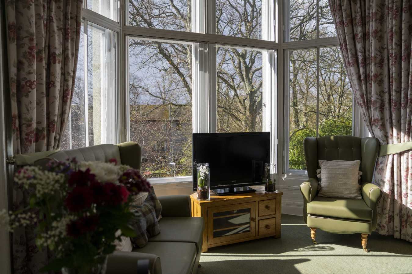 Comfortable care home bedroom lounge featuring a couch, armchair, and TV unit for relaxation. Large windows offer a scenic view of the garden below