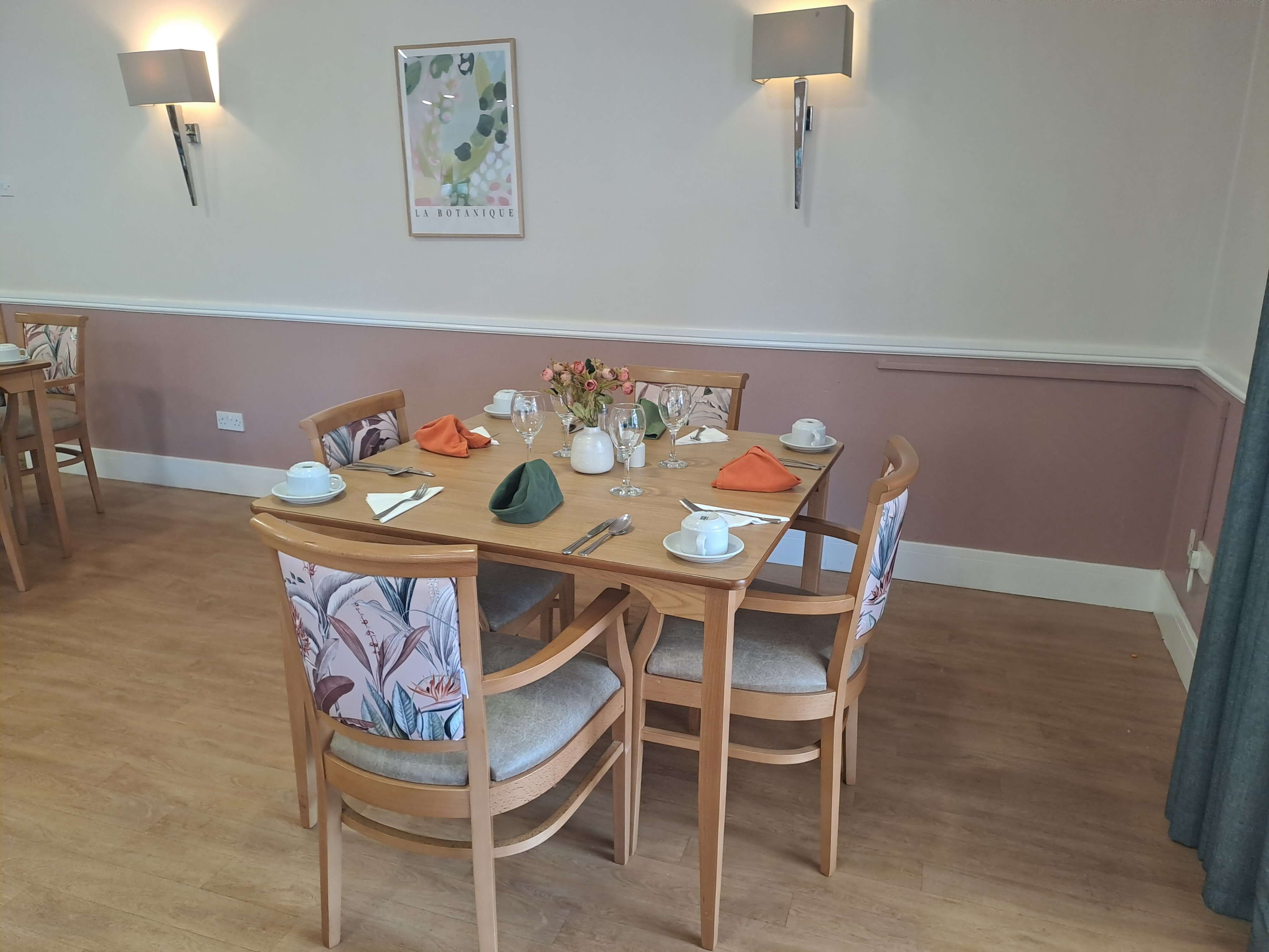Four floral patterned chairs sit around a table in a dining area. The walls are panelled, and the colours are pink and warm. The table is set with four places with two green napkins and two orange ones. There are two ambient wall light fixtures with an abstract artwork in the centre. 
