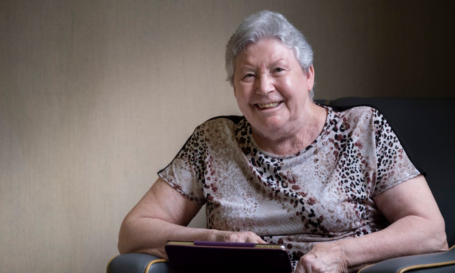 A woman, sitting on a grey chair and smiling, using a tablet 