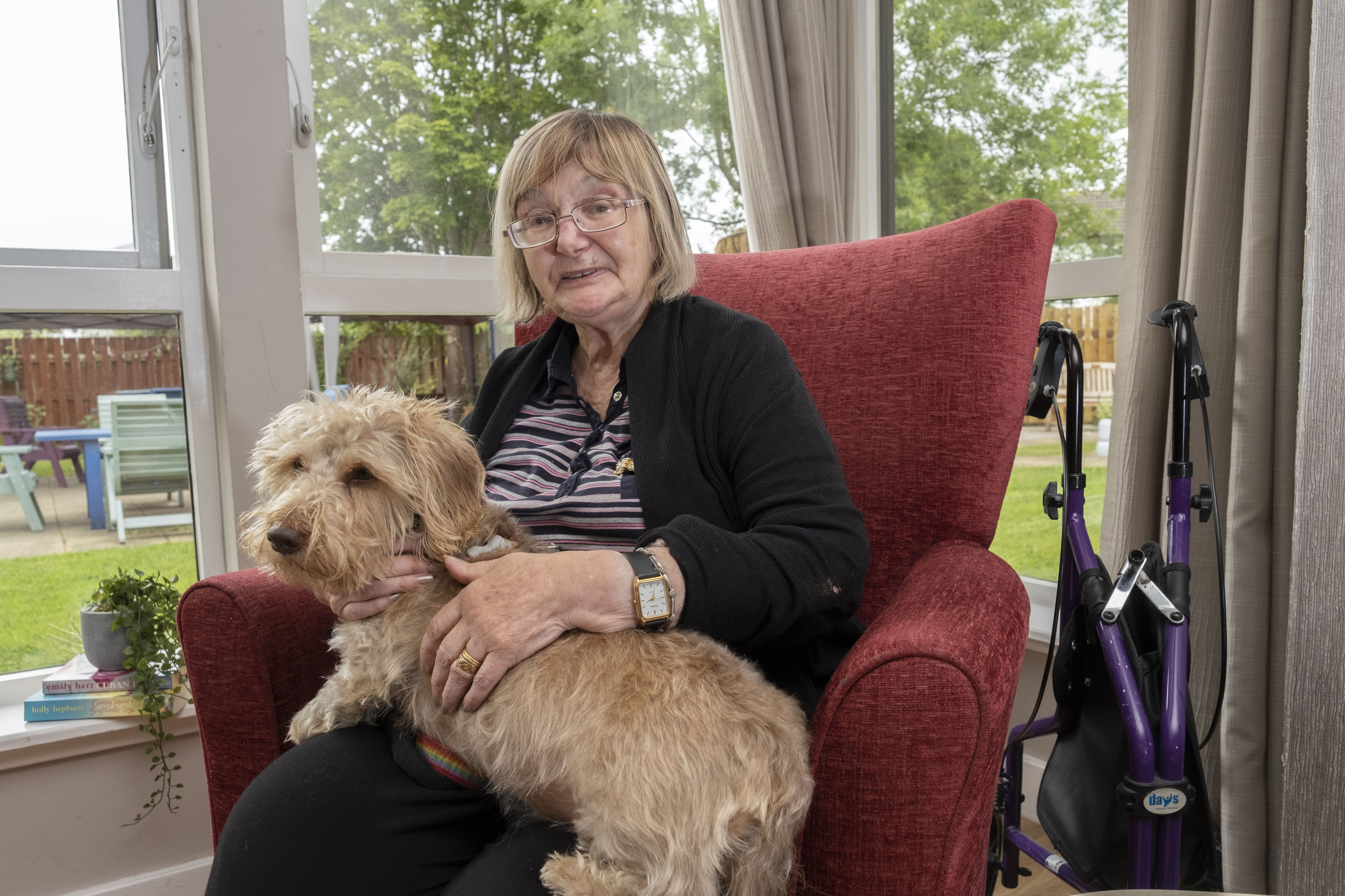 A woman sits on a red armchair next to the window looking out to the garden. She is enjoying a cuddle from therapet, Dexter the dog