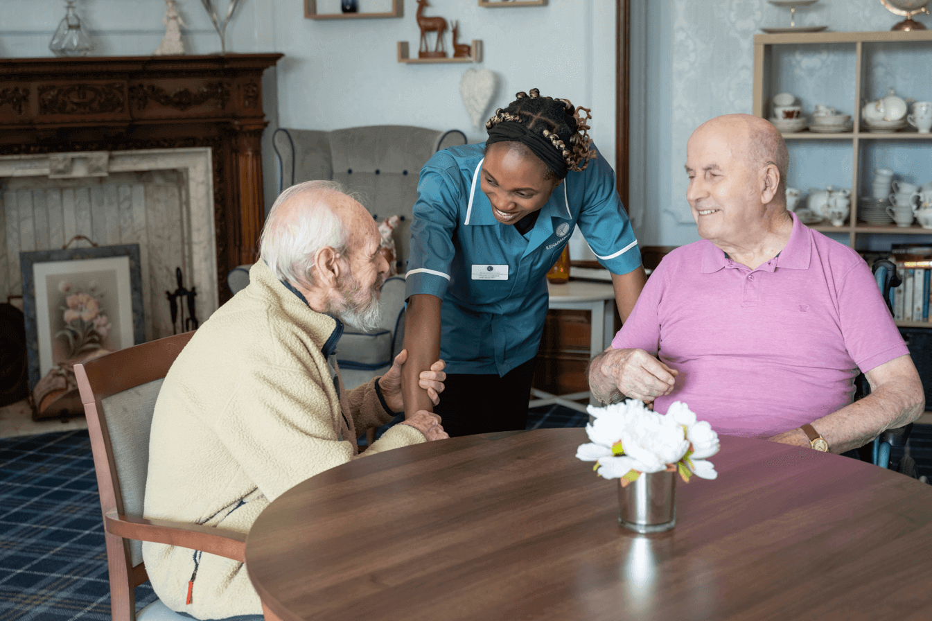 Two residents sit at a wooden table in Cranford Care Homes communal 'Library' space. A staff member attentively chats next to them 