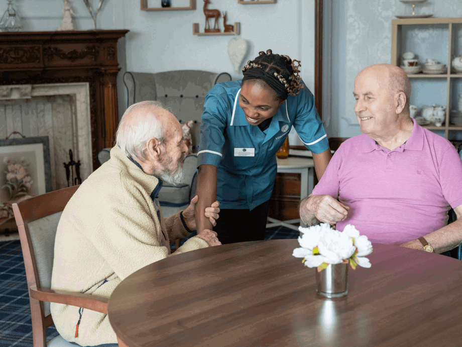 Two residents sit at a wooden table in Cranford Care Homes communal 'Library' space. A staff member attentively chats next to them 