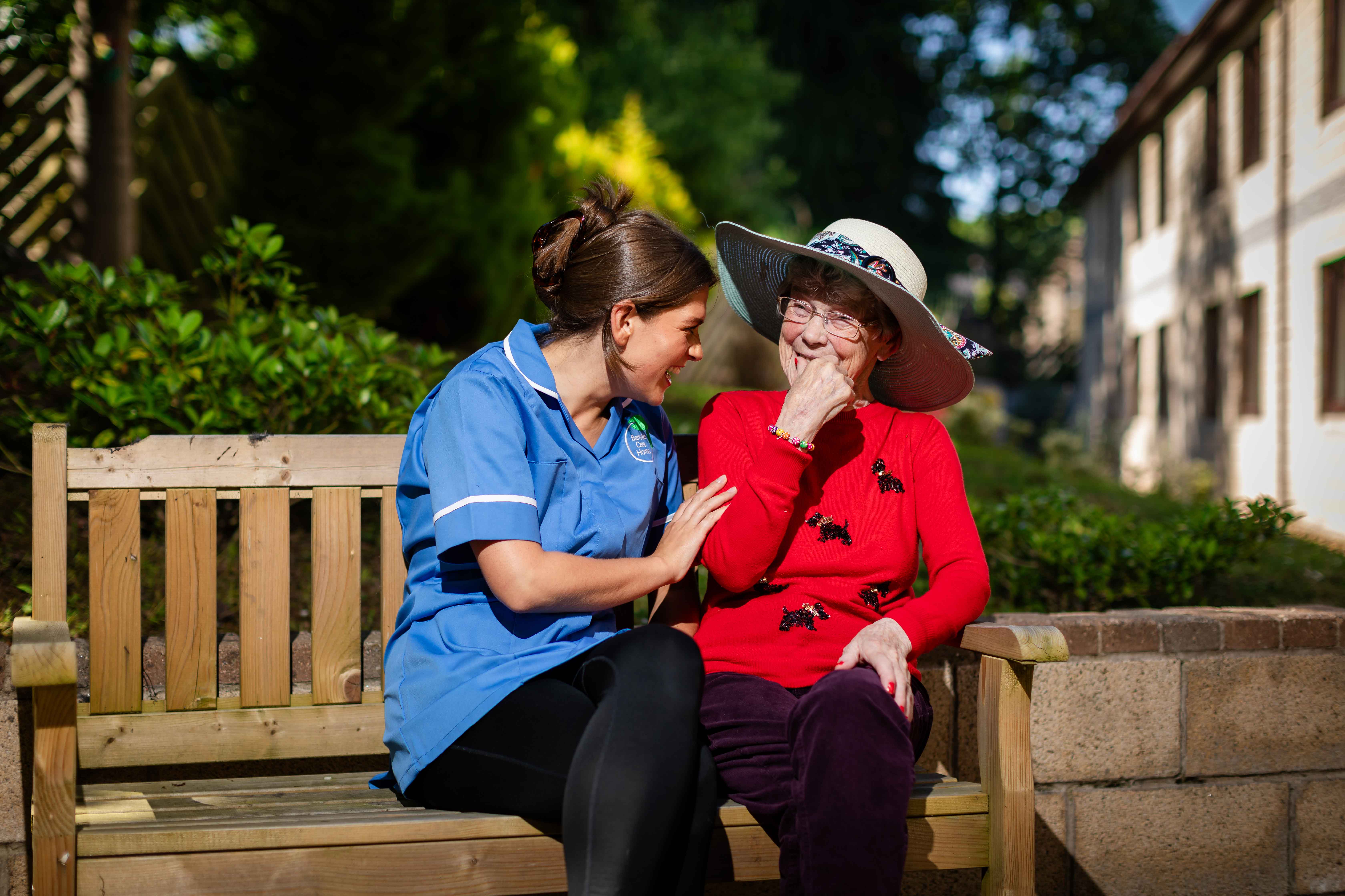 A resident and staff member enjoy one-on-one time outdoors. They sit on a bench in the homes garden area