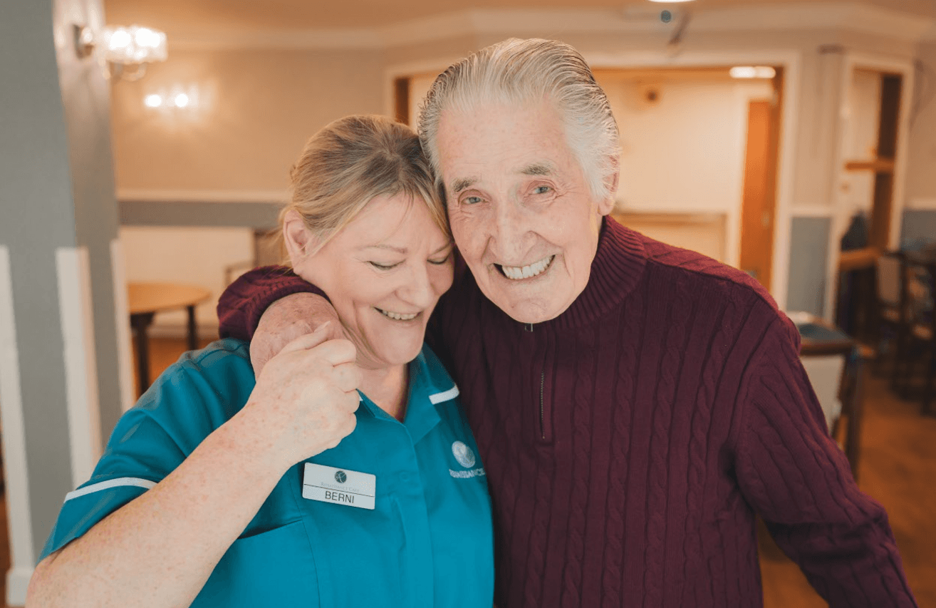 A close-up of a male resident and a female staff member sharing a happy moment. The resident, wearing a maroon sweater, has his arm around the staff member, who is wearing a teal uniform with a name tag that reads "Berni." Both are smiling broadly toward the camera.