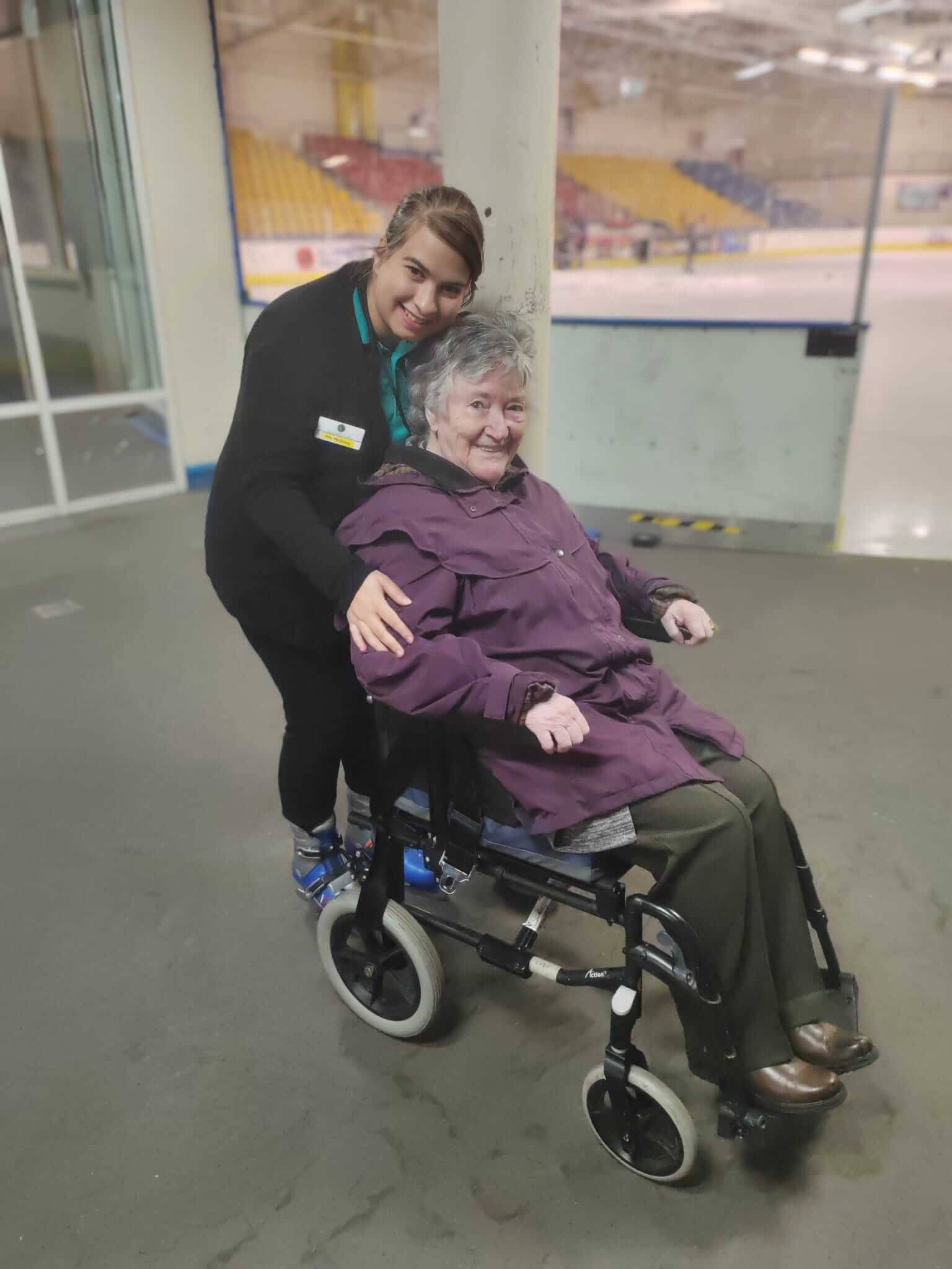 Staff member Ana stands behind resident Lynn, who is seated in a wheelchair eagerly awaiting her turn on the ice rink 
