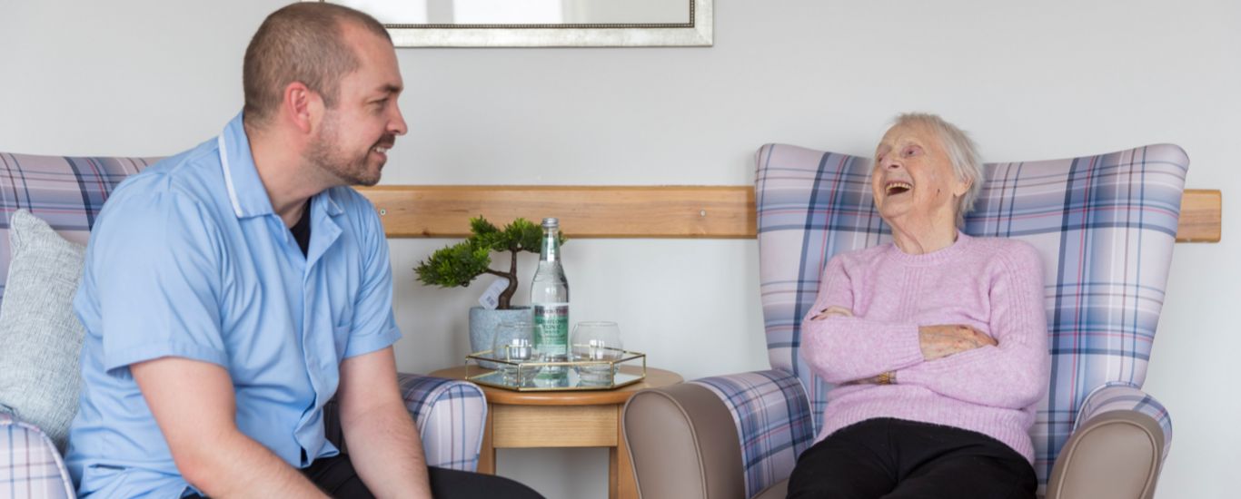 A staff member and resident sit opposite each other enjoying a conversation in a communal lounge 