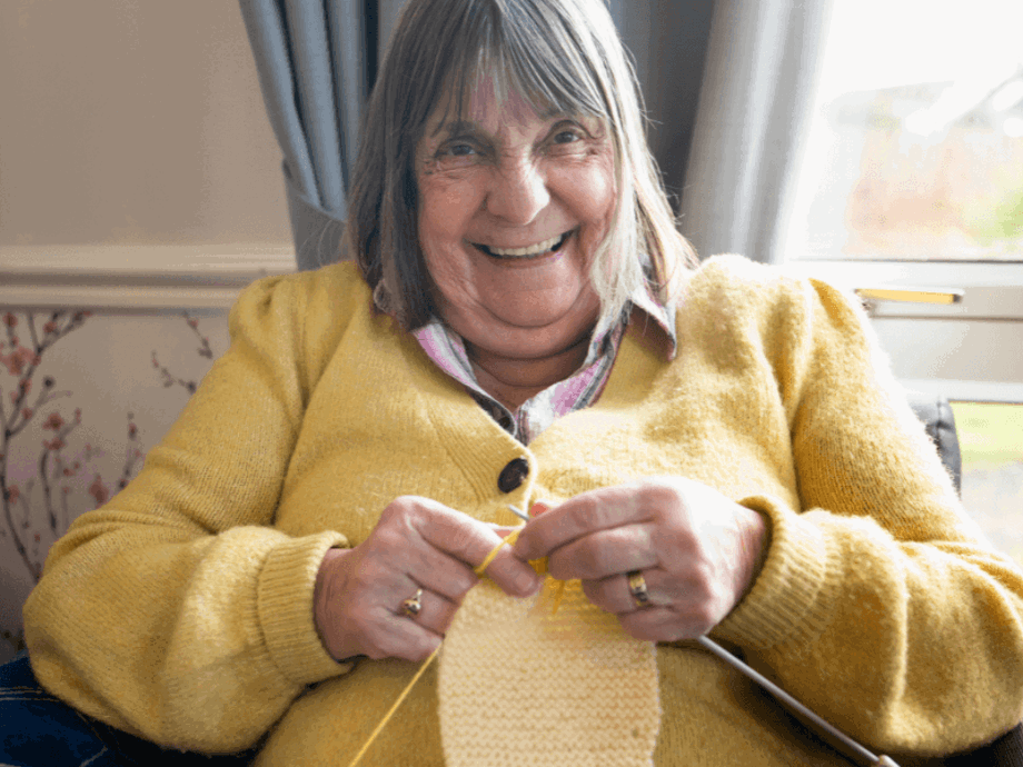 A close-up of an elderly woman with grey-brown hair and a joyful expression, wearing a bright yellow cardigan. She is sitting by a window and actively knitting with yellow yarn.