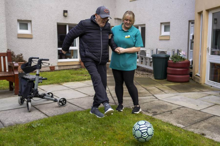 Care home resident stays active with a fun game of football assisted by a caring staff member outdoors
