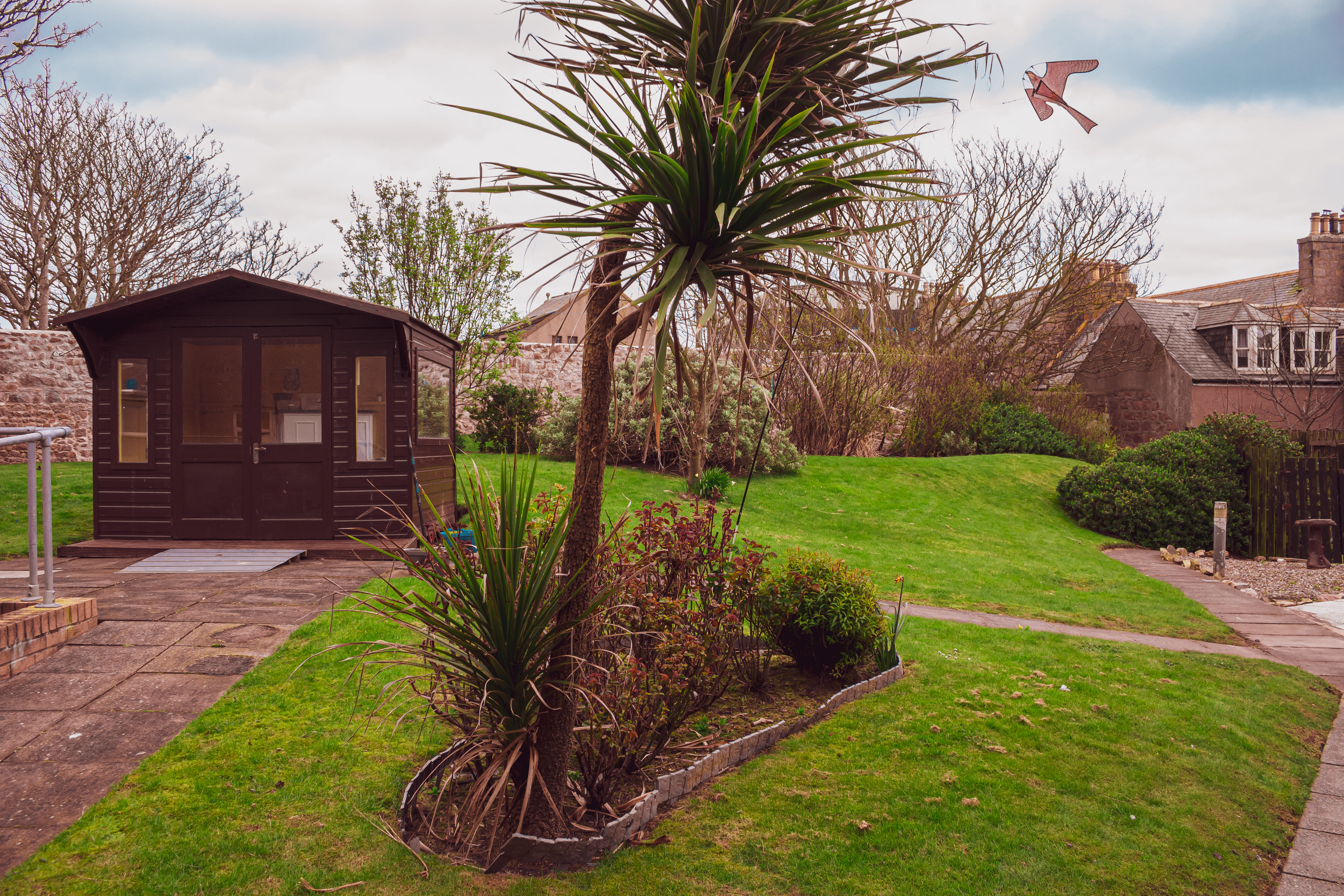 A landscaped garden space with a path and a variety of bushes, hedges & shrubs. There is a large summer house to the left hand side. 