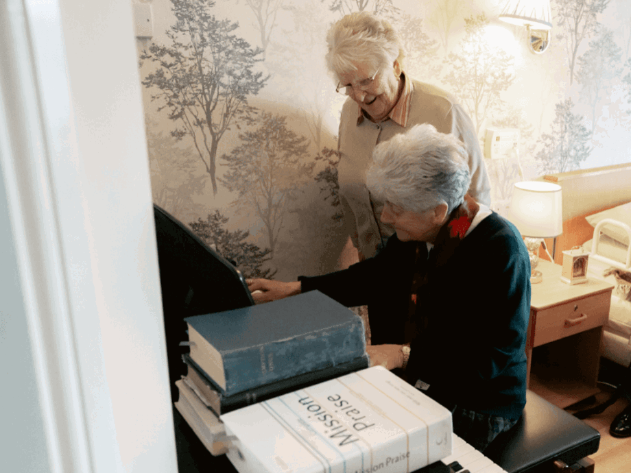Two elderly women are gathered at a piano. One woman is seated and playing the keys, while the other stands beside her, leaning in and smiling. A stack of songbooks, including one titled "Mission Praise," sits on the piano.