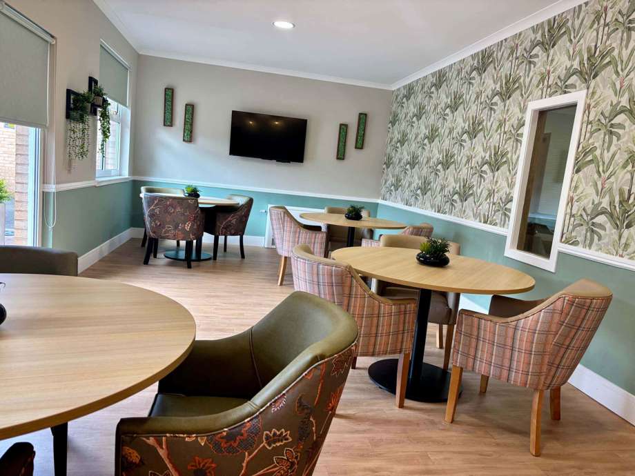 A calm, welcoming garden room space with tables, chairs and greenery scattered throughout, as well as a tv on the back wall. 