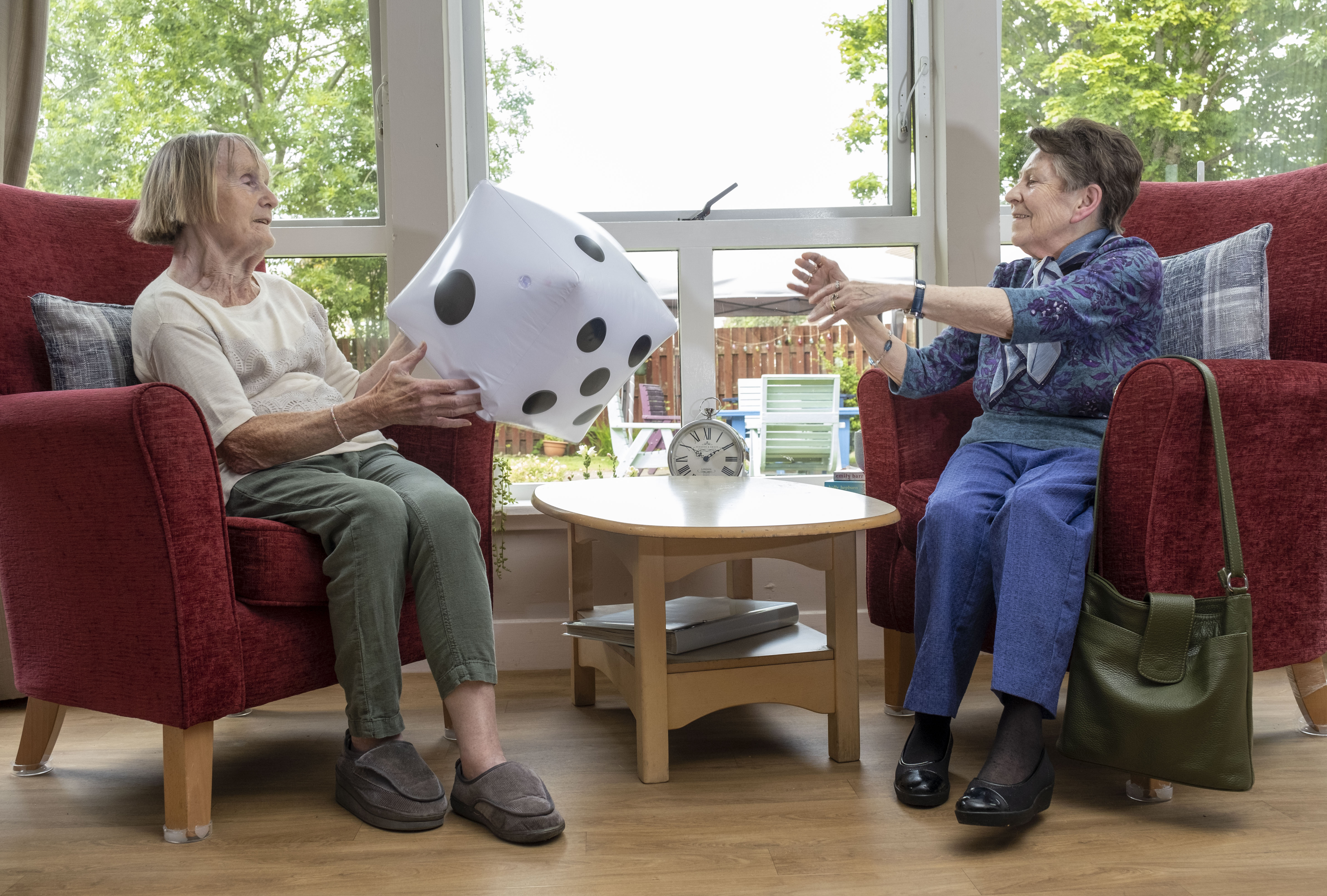 two woman are sitting in a communal lounge area on red armchairs, with a wooden table inbetween. The large windows at the back of them look out into the garden. They are throwing a large inflatable dice to eachother