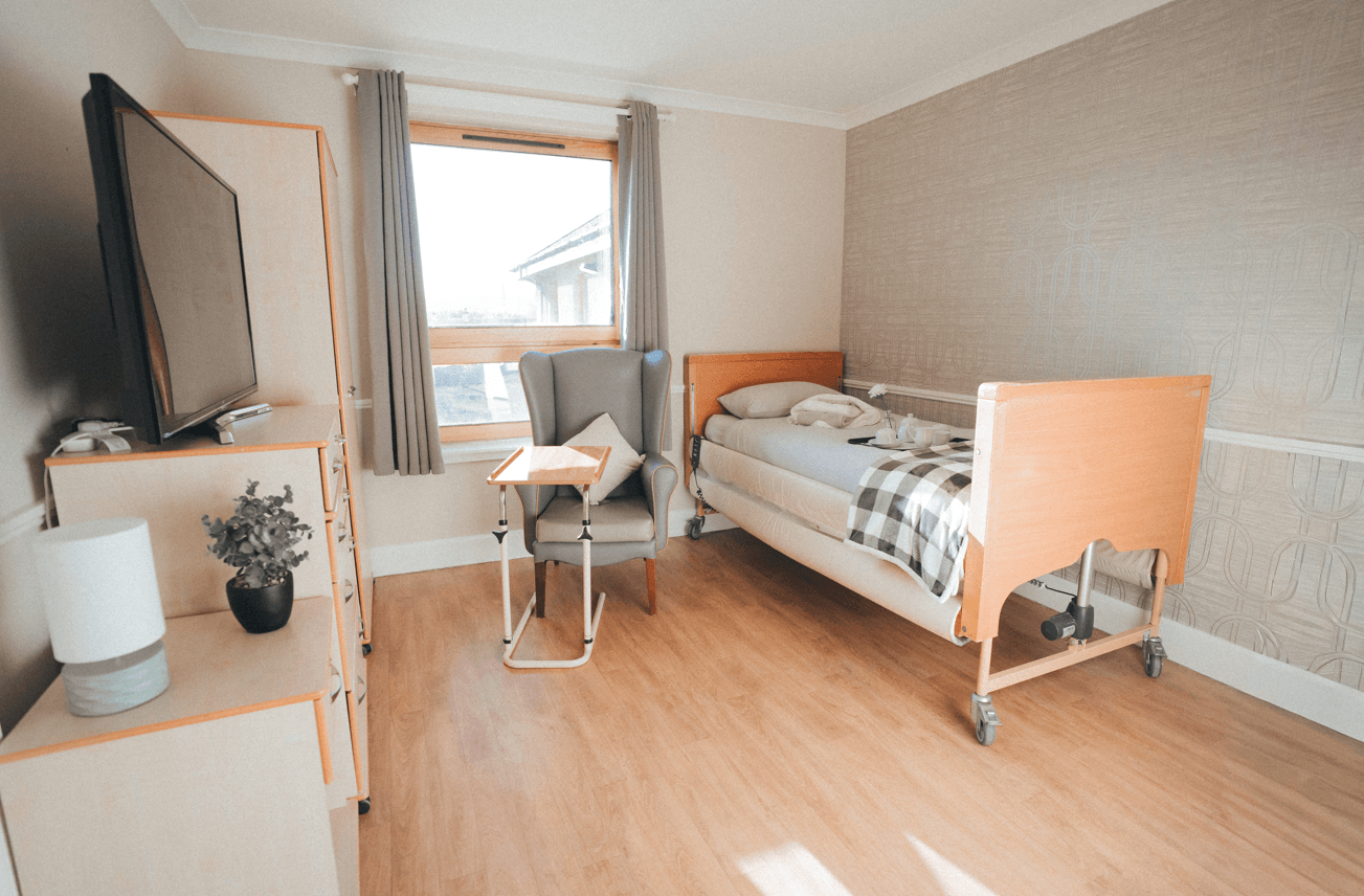 A bright, private bedroom featuring an adjustable bed with wood accents. The room includes a large window, a grey wingback chair with a side table, a television on a light-wood dresser, and neutral-toned wallpaper.