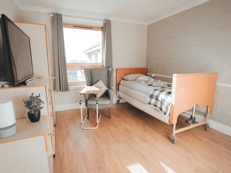 A bright, private bedroom featuring an adjustable bed with wood accents. The room includes a large window, a grey wingback chair with a side table, a television on a light-wood dresser, and neutral-toned wallpaper.