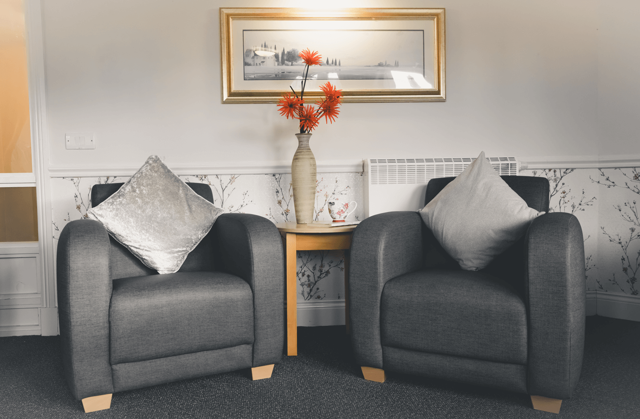 A cosy sitting area featuring two grey upholstered armchairs with silver and grey velvet cushions. Between the chairs is a light wood side table holding a tall, slender vase with bright orange flowers and a floral teacup. A framed landscape painting hangs on the wall above.