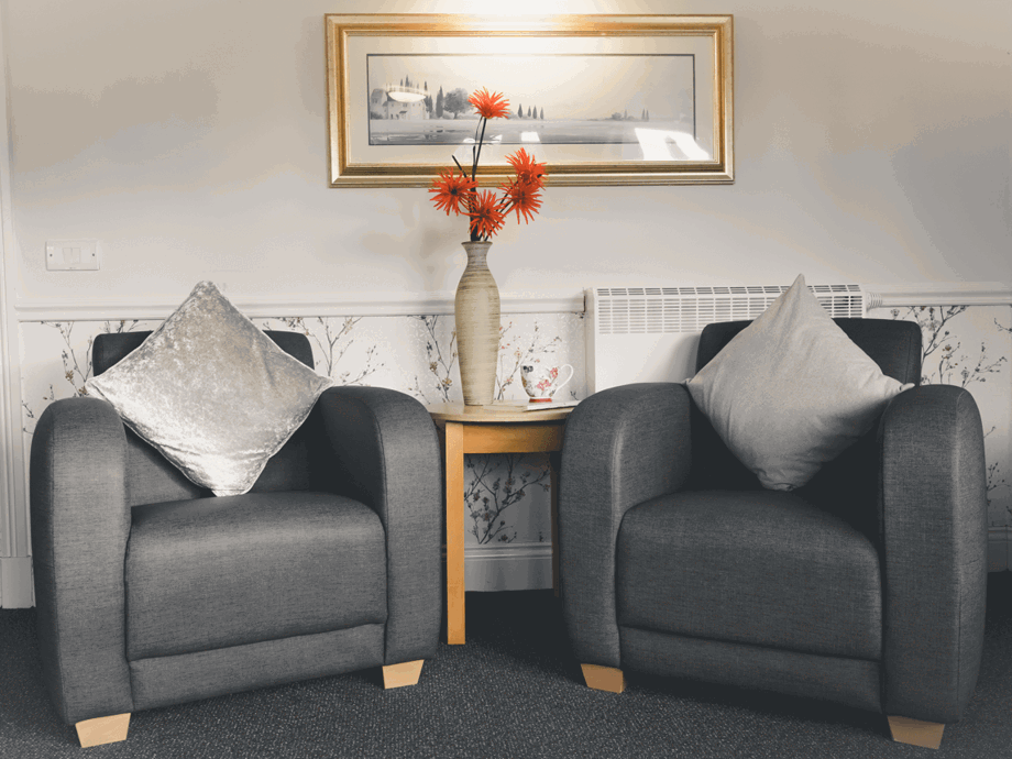 A cosy sitting area featuring two grey upholstered armchairs with silver and grey velvet cushions. Between the chairs is a light wood side table holding a tall, slender vase with bright orange flowers and a floral teacup. A framed landscape painting hangs on the wall above.