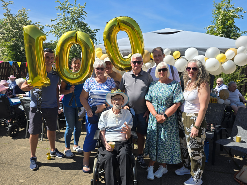 A man is surrounded by his loved ones in Whitecraigs care home garden area as he celebrates his 100th birthday. One person holds three balloons with the mans age. 