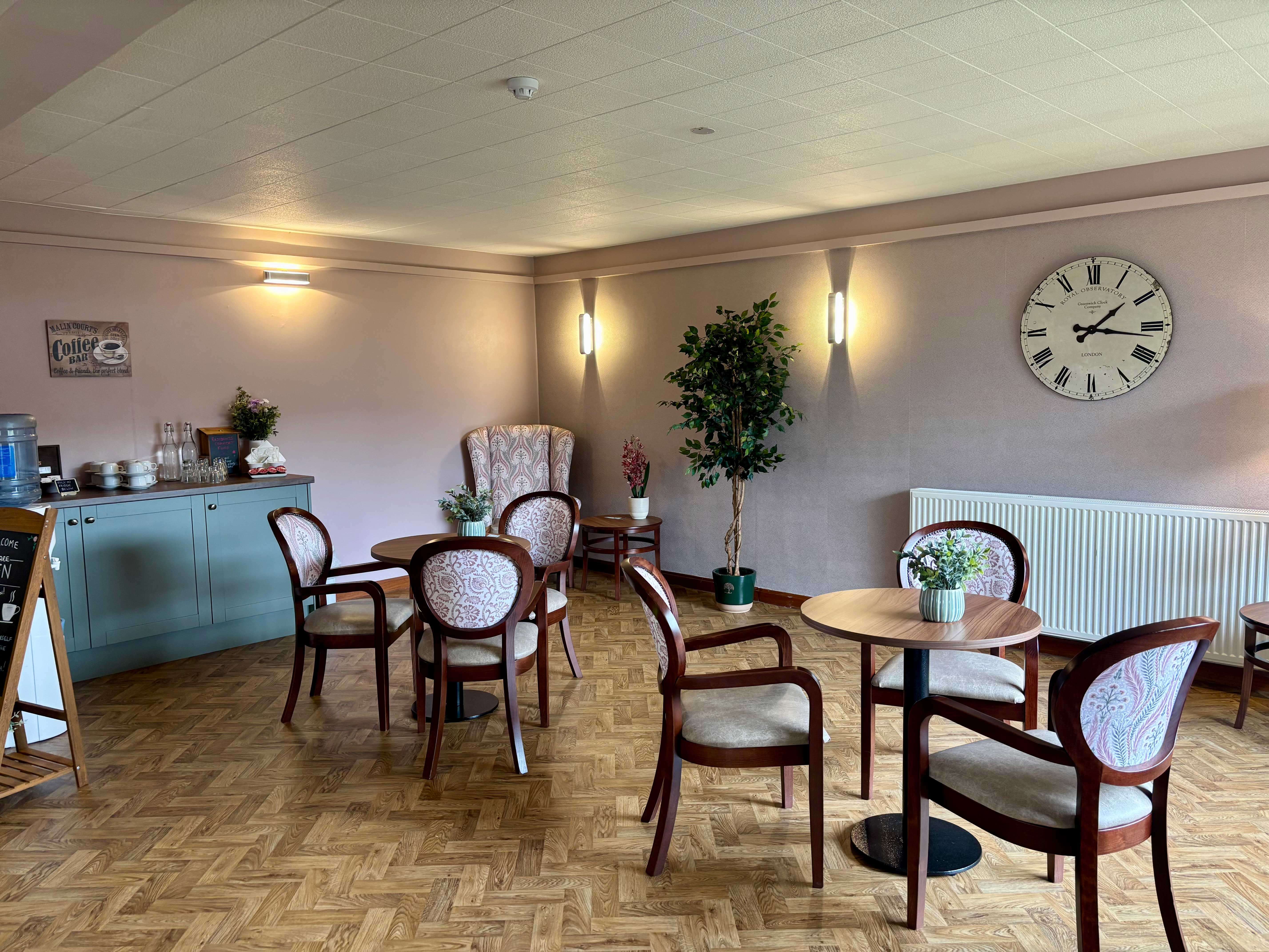 A light and airy cafe area featuring small round tables, wood-framed chairs with light pink patterned upholstery, and soft pink/mauve walls. A large vintage-style wall clock hangs on the right, and a drinks station is on the left.