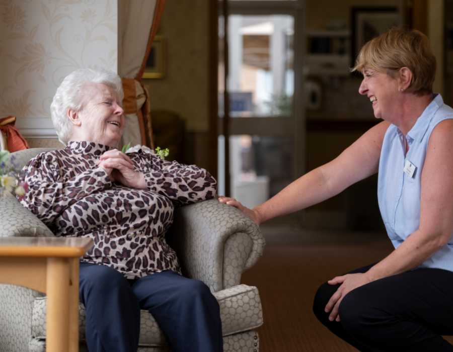 A staff member kneeling down to converse with a woman in a beige chair, they are both smiling and facing one another