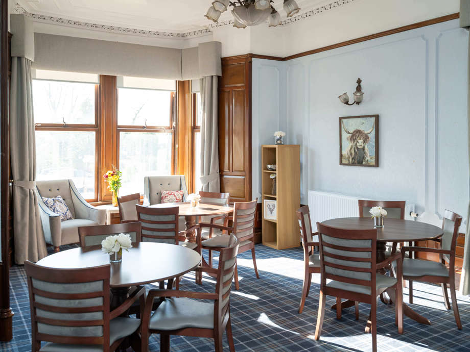 This wide shot captures the lounge area with its arrangement of tables and chairs, emphasising the tall ceilings, detailed cornicing, and prevalent Scottish themes.