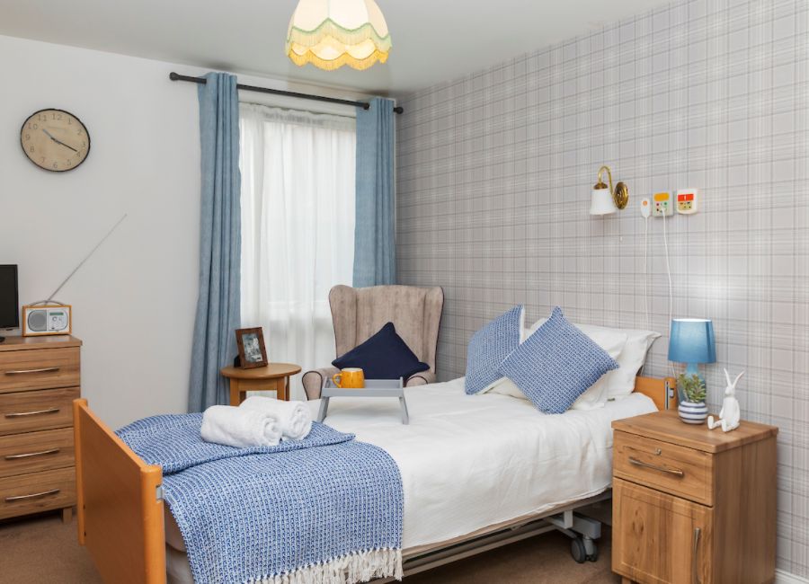 This comfortable bedroom set up compromises of a bedside table, bed, chair, and chest of drawers. The decor is neutral, with pops of blue throughout. There are fluffy towels on the bed and a range of personal belongings giving it a cosy feel