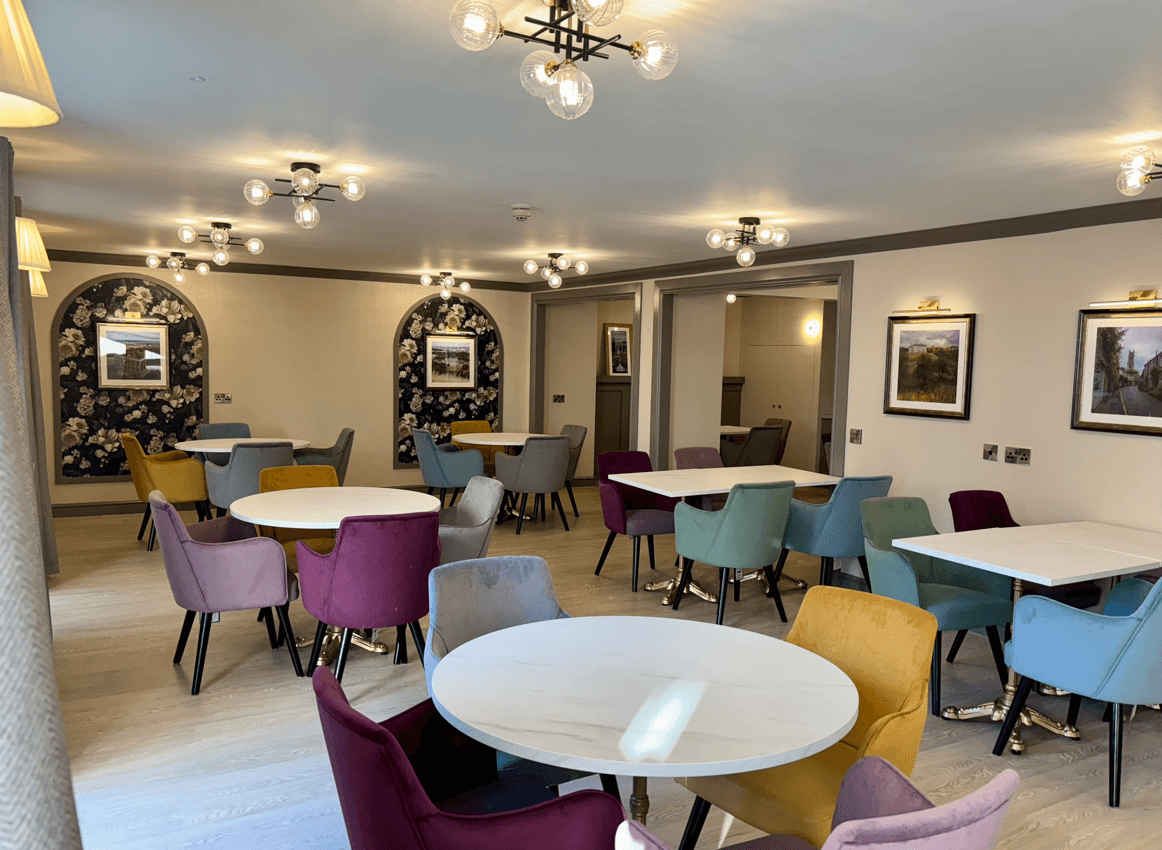 A bright, renovated dining area with multiple round and square white tables. The chairs are upholstered in varied jewel tones like mustard yellow, teal, purple, and grey. The walls are light grey with dark floral arched alcoves and modern ceiling light fixtures.