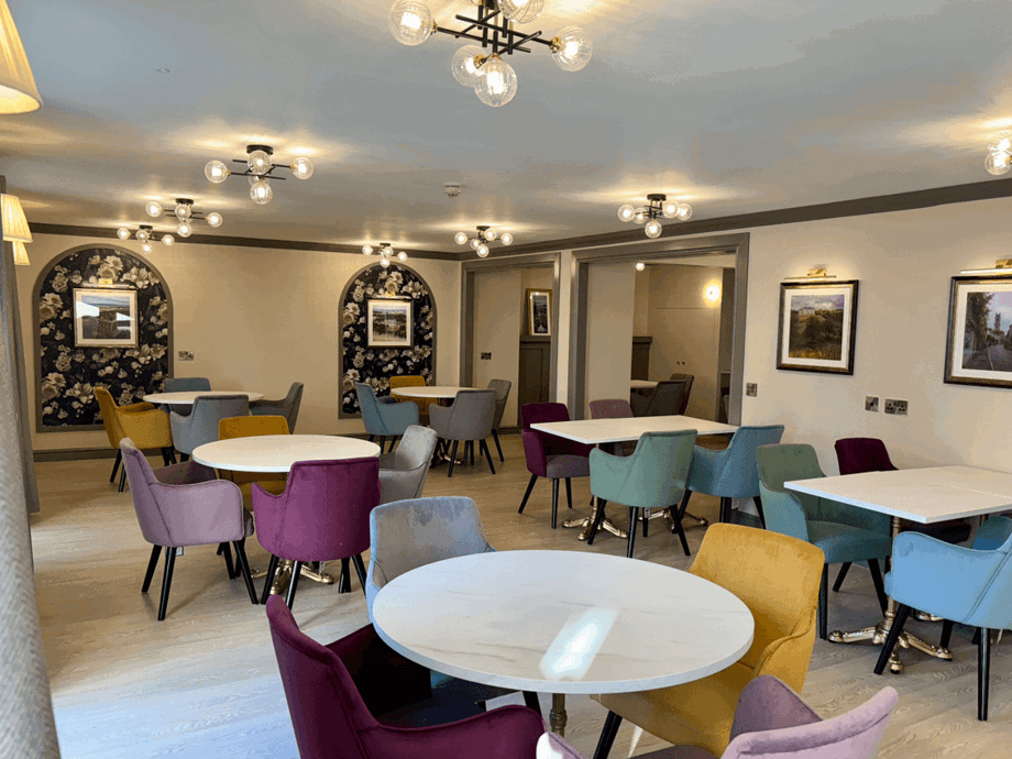 A bright, renovated dining area with multiple round and square white tables. The chairs are upholstered in varied jewel tones like mustard yellow, teal, purple, and grey. The walls are light grey with dark floral arched alcoves and modern ceiling light fixtures.