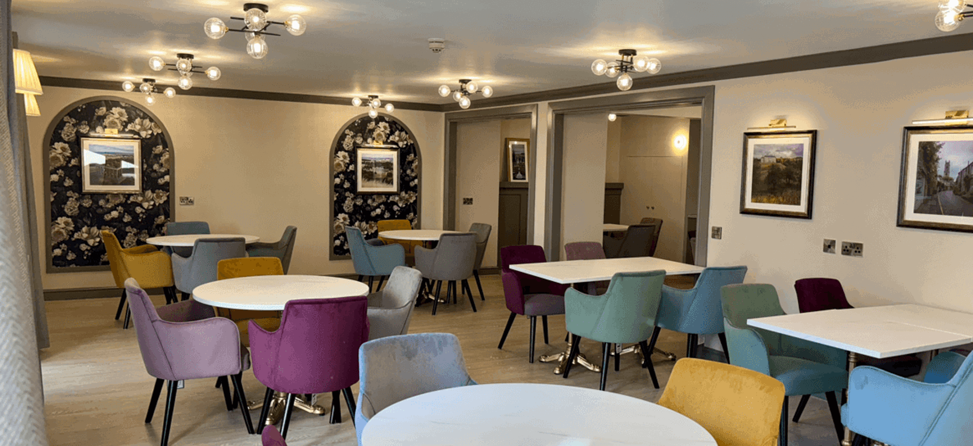 A bright, renovated dining area with multiple round and square white tables. The chairs are upholstered in varied jewel tones like mustard yellow, teal, purple, and grey. The walls are light grey with dark floral arched alcoves and modern ceiling light fixtures.