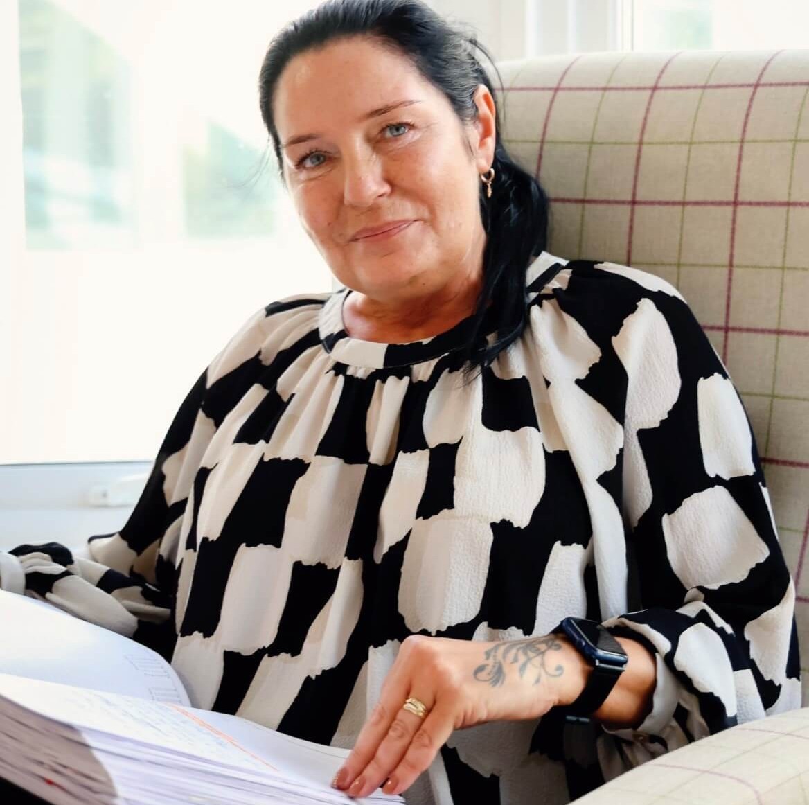 Anna Hoffmann, Care Home Manager, sits on a tartan chair infront of a window. She has shoulder length, dark hair that is tied up and a black and white blouse. 