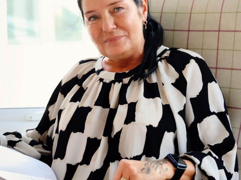 Anna Hoffmann, Care Home Manager, sits on a tartan chair infront of a window. She has shoulder length, dark hair that is tied up and a black and white blouse. 