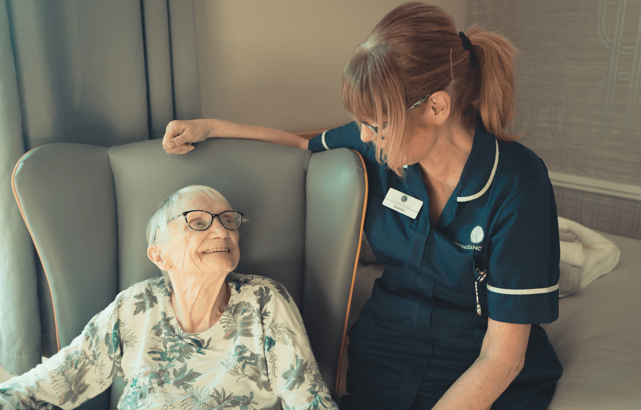 An elderly woman with glasses sits in a grey wingback chair, looking up and smiling at a female caregiver. The caregiver, wearing a dark blue uniform with a name tag that reads "Karen," leans in with a supportive hand on the chair.