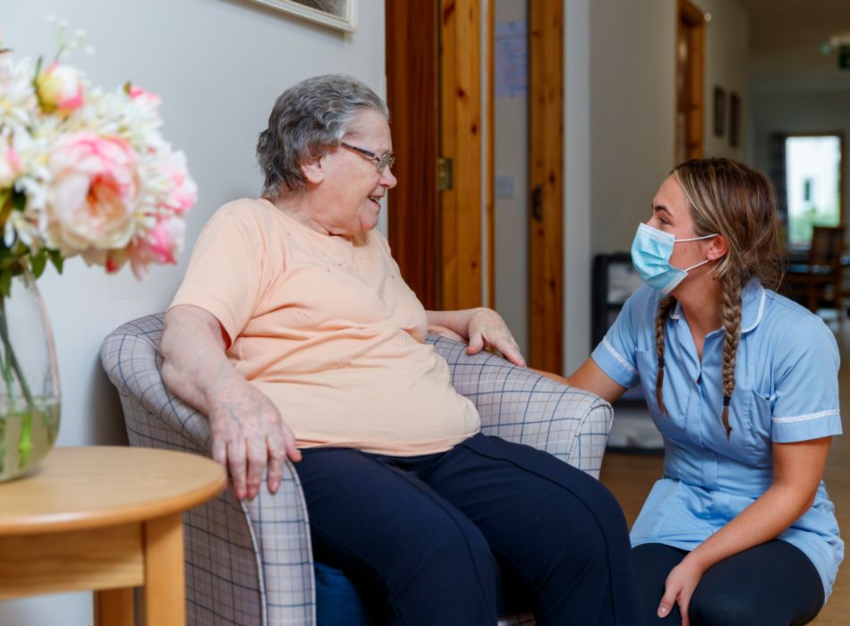 A staff member kneeling down to converse with a resident in a grey tartan chair 