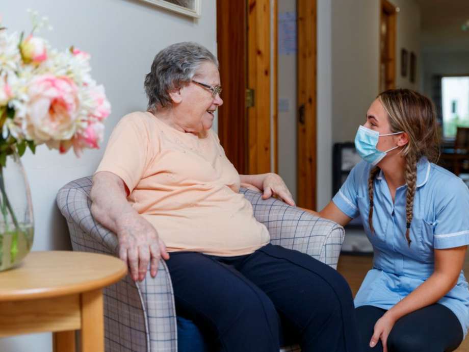 A staff member kneeling down to converse with a resident in a grey tartan chair