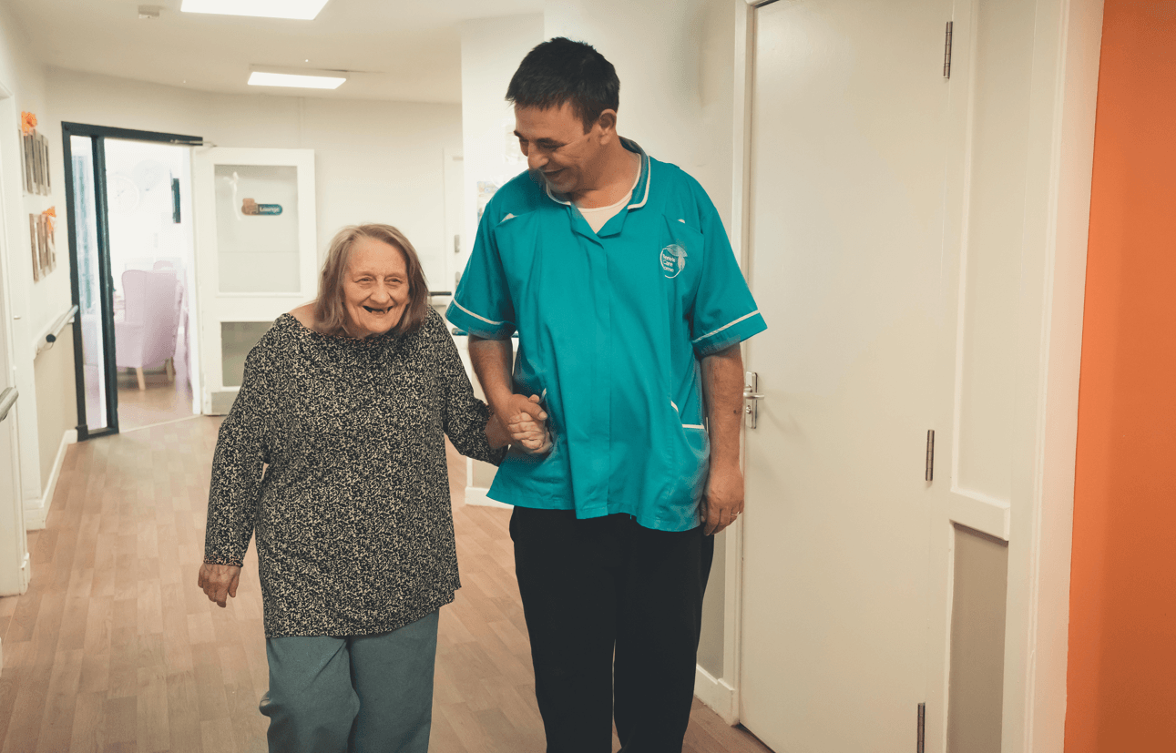 A female resident and a male caregiver walking together down a bright hallway. The resident is smiling broadly while holding the caregiver's hand for support. The caregiver, wearing a teal uniform, looks down at her with a friendly expression