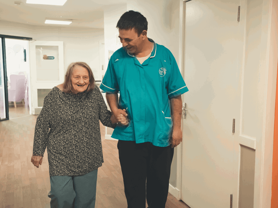 A female resident and a male caregiver walking together down a bright hallway. The resident is smiling broadly while holding the caregiver's hand for support. The caregiver, wearing a teal uniform, looks down at her with a friendly expression