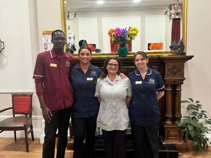 Four smiling staff members in healthcare uniforms standing together in the care homes lounge in front of a fireplace and a large gold-framed mirror.