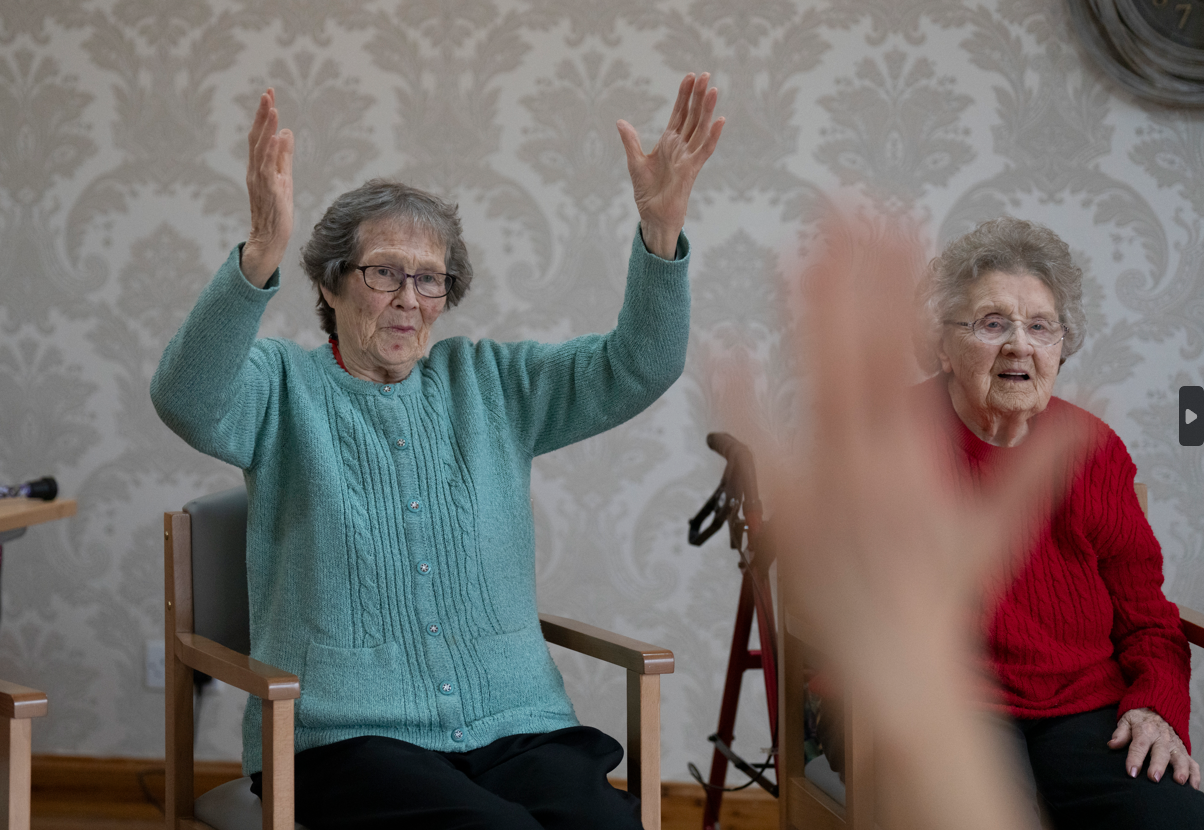 one woman in a blue jumper and one in a red knit copying a yoga instructors movements, their hands are in the air
