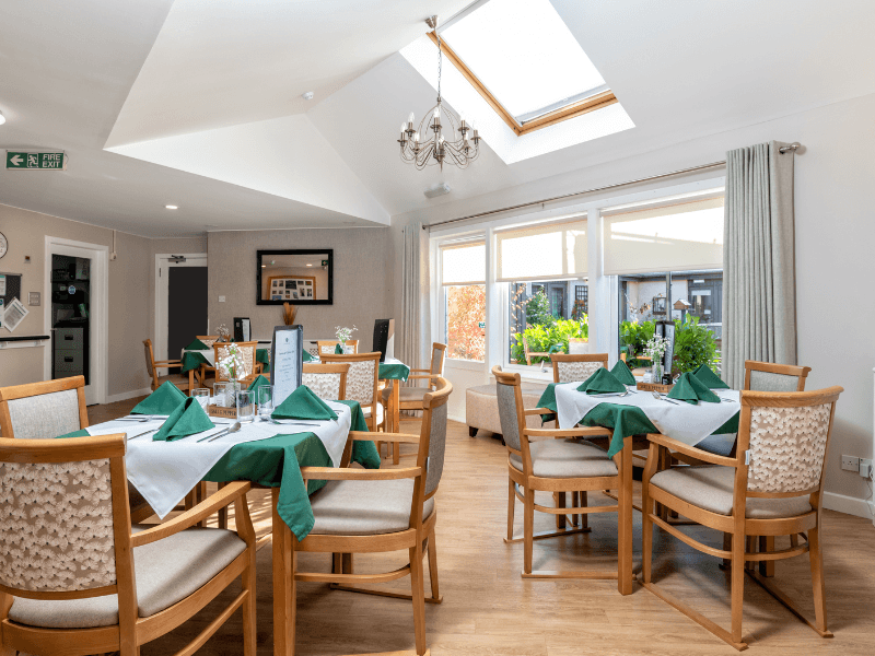 A bright, airy dining room featuring a large skylight and a chandelier. Several wooden dining tables are neatly set with white linens, green napkins, and small floral centerpieces. Large windows and a glass door look out onto a lush green garden.