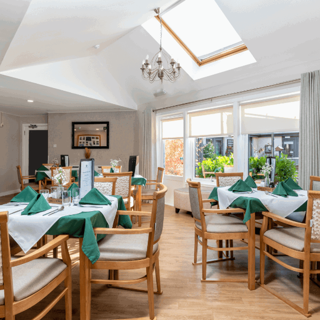 A bright, airy dining room featuring a large skylight and a chandelier. Several wooden dining tables are neatly set with white linens, green napkins, and small floral centerpieces. Large windows and a glass door look out onto a lush green garden.