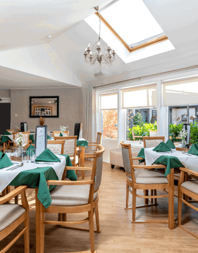 A bright, airy dining room featuring a large skylight and a chandelier. Several wooden dining tables are neatly set with white linens, green napkins, and small floral centerpieces. Large windows and a glass door look out onto a lush green garden.