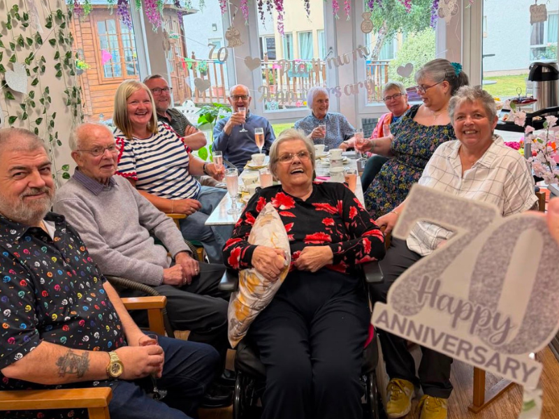 A family of 10 gather around a large table, celebrating Doug and Doris' anniversary. There are flowers and decorations on the walls, and large windows to the back show a view into the garden. 