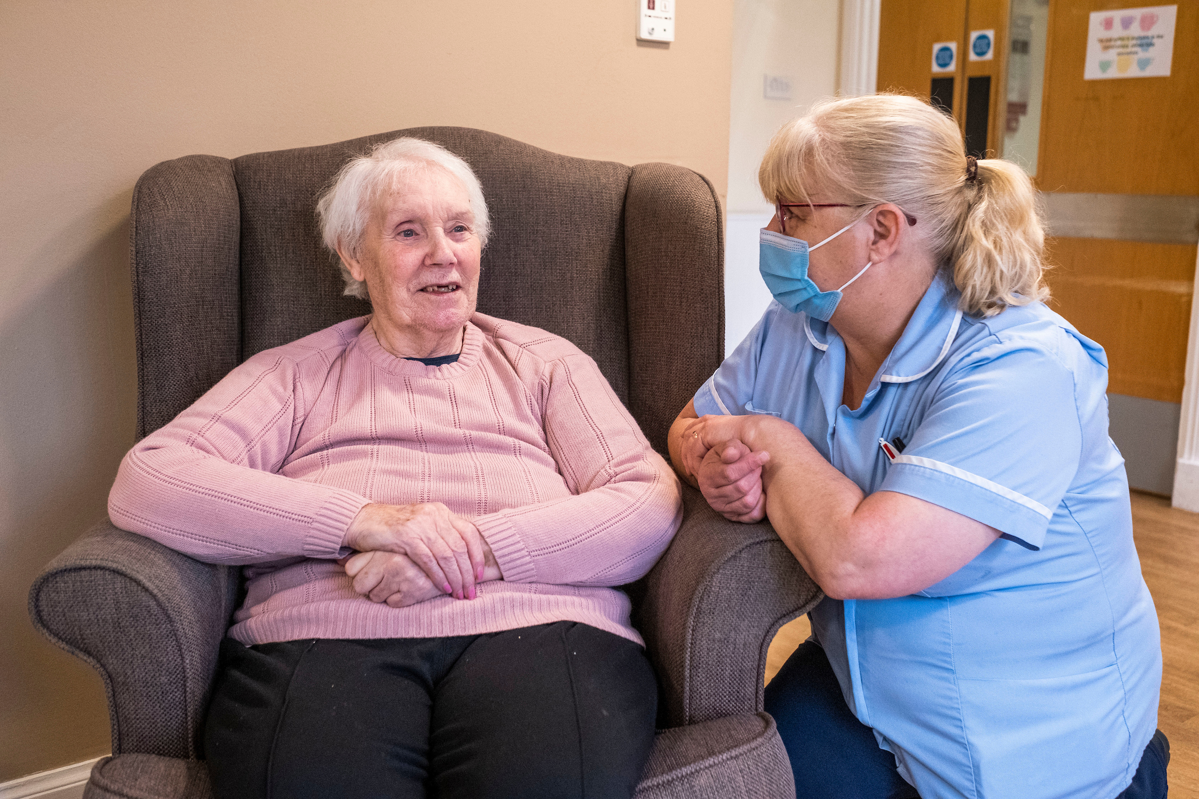 Caregiver kneels to talk attentively with a woman seated in a grey chair