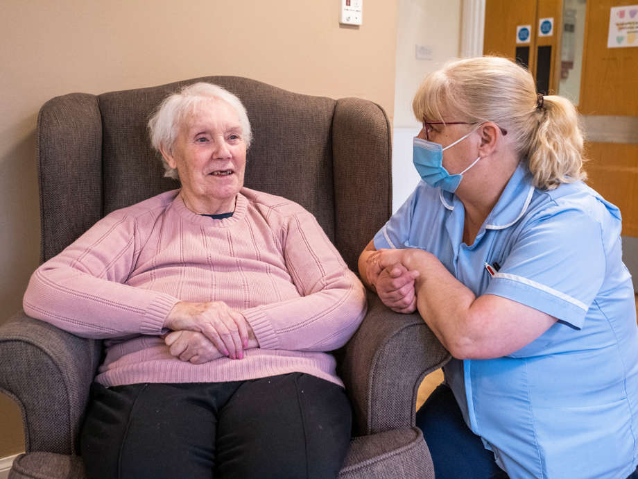 Caregiver kneels to talk attentively with a woman seated in a grey chair