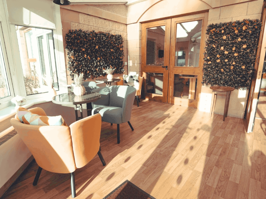 A sunny sitting area featuring orange and blue upholstered chairs around small glass-topped tables. Two large decorative floral wall panels add texture to the stone walls.