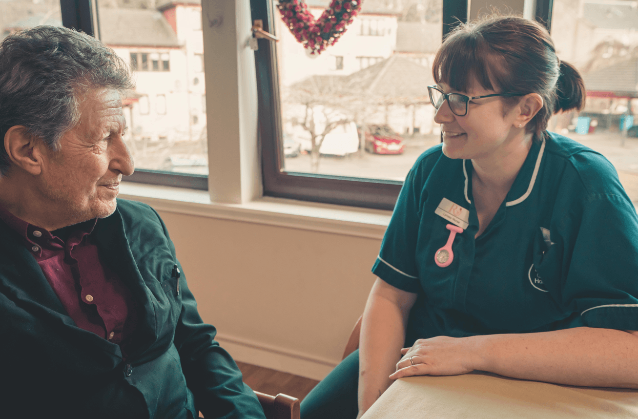 A close-up of an elderly man and a female nurse sitting at a table together. The nurse, wearing a dark green uniform and glasses, is smiling warmly as she listens to the man. They are positioned in front of a large window overlooking a residential area.