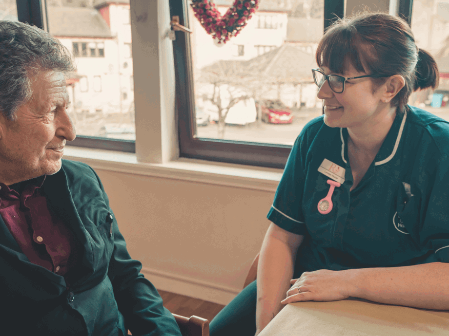 A close-up of an elderly man and a female nurse sitting at a table together. The nurse, wearing a dark green uniform and glasses, is smiling warmly as she listens to the man. They are positioned in front of a large window overlooking a residential area.