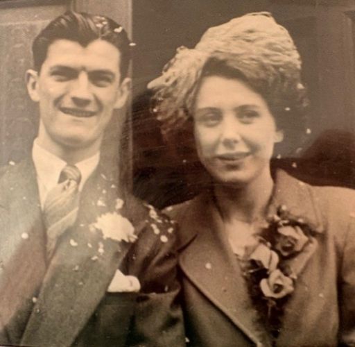 A black-and-white vintage photograph of a young couple on their wedding day. The man is on the left, wearing a dark suit with a white carnation boutonniere and a patterned tie, his hair neatly styled. The woman is on the right, wearing a structured coat with a large floral corsage and an ornate, feathered or textured fascinator hat. Confetti is visible scattered across their clothing.