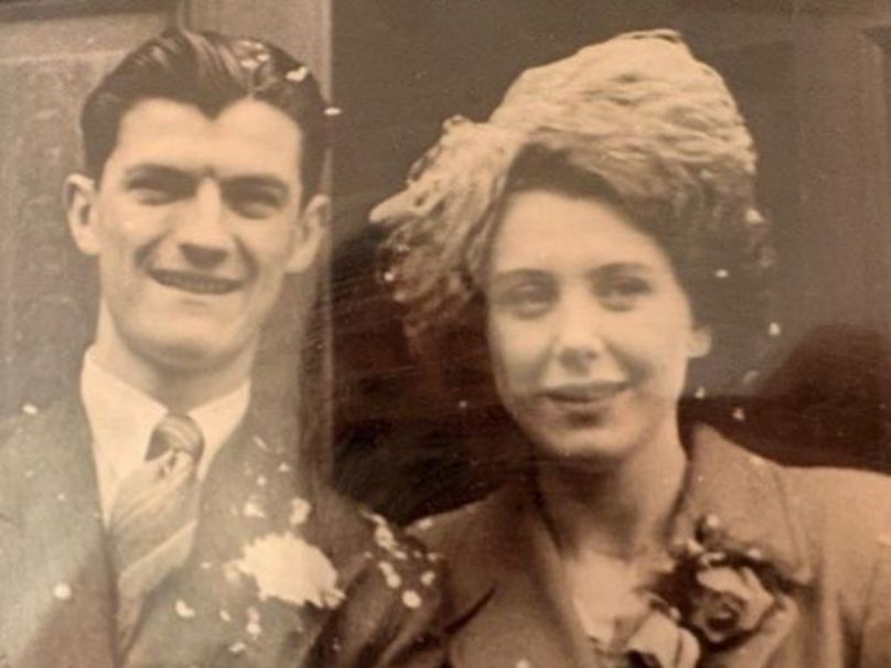 A black-and-white vintage photograph of a young couple on their wedding day. The man is on the left, wearing a dark suit with a white carnation boutonniere and a patterned tie, his hair neatly styled. The woman is on the right, wearing a structured coat with a large floral corsage and an ornate, feathered or textured fascinator hat. Confetti is visible scattered across their clothing.
